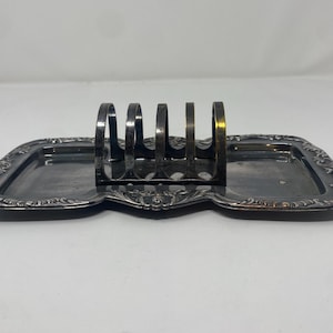 May include: An antique silver-toned toast rack with a decorative, ornate design. The rack features a rectangular base with a central section for the toast slots. The edges of the base are detailed with floral patterns. The rack is designed to hold multiple slices of toast.
