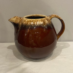 Puede incluir: Una jarra de cerámica esmaltada marrón con un diseño de esmalte blanco y marrón. La jarra tiene un cuerpo redondo y un asa.