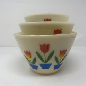 May include: Three off-white mixing bowls stacked together. Each bowl has a tulip design with red and yellow flowers in blue pots. The bowls are made of a durable material. The bowls are measured in centimeters.