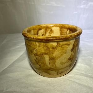 May include: A small, round ceramic bowl with a speckled, mottled pattern in shades of tan and brown. The rim is a darker brown. The bowl has a rustic, handmade appearance, suitable for serving or display.
