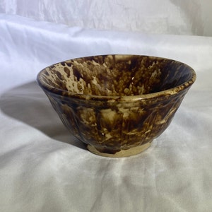 Puede incluir: Un tazón de cerámica esmaltado en marrón y crema con un patrón moteado. El tazón tiene una forma redondeada y un borde ligeramente acampanado.