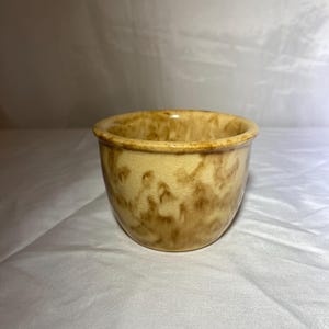 Puede incluir: Un pequeño cuenco redondo de cerámica con una base de color amarillo claro y un patrón moteado marrón. El cuenco tiene una forma simple y redondeada y un acabado liso y brillante. El interior es de color amarillo claro sólido.