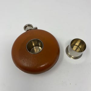 Könnte beinhalten: Eine braune, lederbezogene Flasche mit silbernem Rand und einem passenden Silberbecher. Die Flasche hat eine runde Form mit eingebautem Becherhalter. Der Becher ist separat.