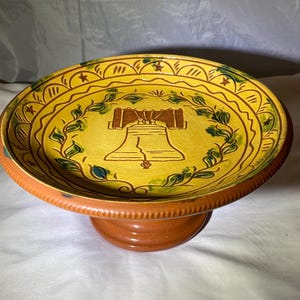 May include: A yellow and brown ceramic pedestal dish with a painted design of the Liberty Bell surrounded by green leaves. The dish has a scalloped edge and a brown base.