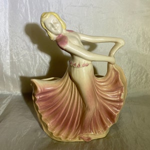 Puede incluir: Un macetero de cerámica con forma de mujer en un vestido rosa fluido. La mujer tiene el pelo rubio y una expresión facial delicada. El macetero es una pieza vintage con un diseño único.