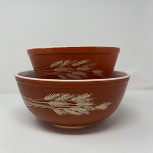 Puede incluir: Dos cuencos para mezclar anidados, de color óxido, con un diseño de espiga de trigo. Los cuencos tienen un borde blanco y un patrón de trigo beige. El cuenco más grande está en la parte inferior y el cuenco más pequeño está anidado dentro.
