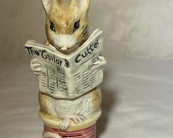 May include: A ceramic figurine of a mouse reading a newspaper. The mouse is tan and white, with black eyes and whiskers. The newspaper reads "The Tailor" and "Cutter". The figurine is on a spool of thread.