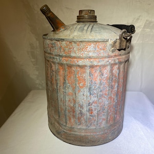 Puede incluir: Un antiguo bidón de gasolina metálico con un aspecto desgastado y envejecido. El bidón es cilíndrico con un pico, una tapa de rosca y un asa negra. El exterior presenta una mezcla de pintura naranja y gris, mostrando signos de uso.