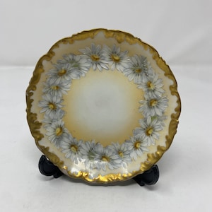 Puede incluir: Un plato pequeño y decorativo con borde festoneado y borde dorado. El plato presenta un anillo de margaritas blancas con centros amarillos alrededor del borde. El centro del plato es de color amarillo claro.