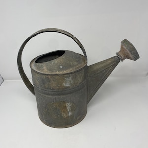 May include: A weathered gray metal watering can featuring a cylindrical body, a large arched handle attached to the top, and a tapered spout with a detachable rose head. The surface shows signs of oxidation and discoloration throughout the exterior.