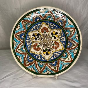 Puede incluir: Un plato de cerámica decorativo con un diseño floral detallado. El plato presenta un patrón central de estrella en amarillo, negro y rojo, rodeado por un borde floral azul y turquesa. El borde es verde y el plato se exhibe sobre un soporte transparente.