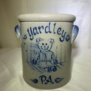 Puede incluir: Un tarro de gres con detalles pintados en azul. El tarro presenta la palabra "yardley" en escritura, una ilustración de oso de peluche y las letras "PA". Tiene dos asas pequeñas y un borde redondeado.