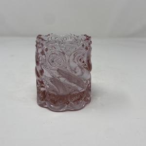 May include: A cylindrical glass votive holder in a soft pink hue, featuring a raised, textured pattern of swirling scrolls and circular motifs that wrap around the exterior. The top and bottom edges have a scalloped, beaded design.