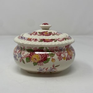 May include: A cream-colored ceramic sugar bowl with a matching lid, featuring a colorful floral pattern of red, yellow, and blue flowers with green leaves. The bowl has a rounded shape with a scalloped rim and a small knob on the lid.