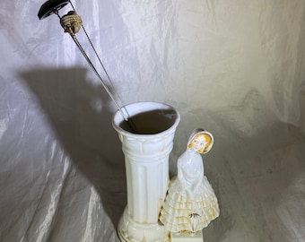 May include: White ceramic vase with a column design and a figurine of a girl in a white dress holding an umbrella. The vase has a green and black pin holder.