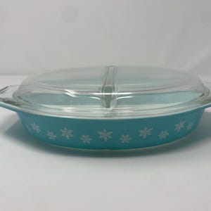 May include: A turquoise Pyrex divided casserole dish with a clear glass lid. The dish features a snowflake pattern around the exterior. The lid is divided into two sections. The dish has two handles.
