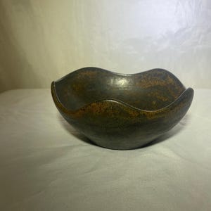 Puede incluir: Un cuenco de cerámica oscura hecho a mano con un borde ondulado único. El cuenco tiene una superficie texturizada con toques de oro y marrón, creando una estética rústica y artística. El cuenco está diseñado para uso decorativo o funcional.