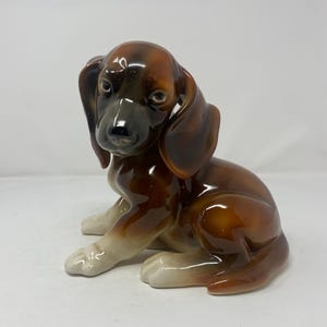 以下が含まれることがあります： 長い垂れ耳をした子犬が座っている姿の光沢のある陶器製の置物。頭部と耳に濃い茶色の陰影がある豊かな茶色の毛並みが特徴で、明るいクリーム色の足と滑らかで反射する仕上げが対照的です。