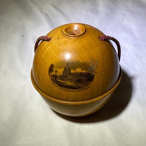 May include: A wooden ball with a string handle, featuring a black and white image of the United States Capitol building in Washington, D.C. The ball is painted with a light brown stain and has a small hole at the top.