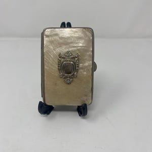 Puede incluir: Un pequeño compacto rectangular antiguo con acabado de nácar. El compacto presenta un emblema decorativo plateado en la parte delantera y está sostenido por un soporte negro. El compacto está cerrado.