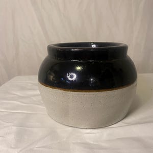 May include: A small, round ceramic jar with a dark navy blue top and a light beige bottom. A thin brown band separates the two colors. The jar has a wide opening and a smooth, glossy finish.