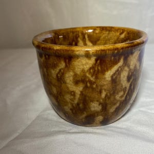 Puede incluir: Un pequeño cuenco de cerámica redondo con un esmalte moteado marrón y beige. El cuenco tiene un borde ligeramente ensanchado y un acabado liso y brillante. El interior es de un tono marrón más claro.