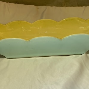 Puede incluir: Un macetero de cerámica azul claro y amarillo con un borde festoneado. El macetero está diseñado con un patrón ondulado en forma de hoja.