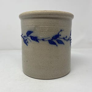 May include: A cylindrical beige stoneware crock with a speckled finish. It features a blue painted vine pattern encircling the middle and a stamped rectangular logo that reads Salmon Falls Stoneware 1995.