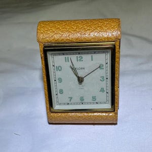 May include: A vintage tan leather-covered alarm clock with a white face and black hands. The clock face has green numbers and the word "FLORN" printed on it.