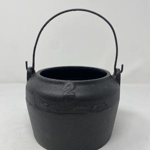 May include: A black cast iron pot with a textured surface and a rounded body. The pot features a thin, arched metal handle attached to two side lugs. The number 2 is embossed on the upper rim of the pot, which has a wide, open circular top.