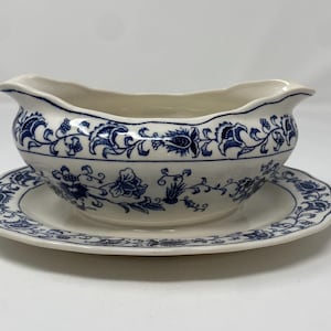 May include: Cream-colored gravy boat and saucer set with blue floral patterns. The gravy boat has a scalloped rim and a handle. The saucer is round with a matching blue floral design. A classic addition to any dining table.