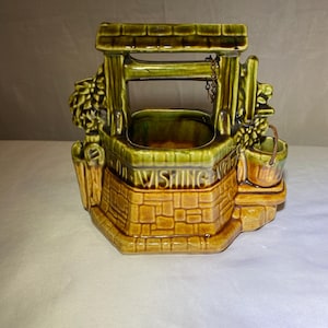 Puede incluir: Una maceta de cerámica con forma de pozo de los deseos, con techo y detalles verdes, base de ladrillo marrón y la palabra "Wishing" en la parte delantera. El pozo tiene un pequeño cubo colgando a un lado y follaje decorativo.