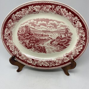 May include: An oval ceramic platter featuring a deep red transferware pattern on a cream background. The center depicts a suspension bridge over a river with trees and clouds. The wide border displays a detailed repeating leaf and floral motif.