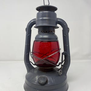 May include: A dark gray metal hurricane lantern featuring a vibrant red glass globe. The lantern has a sturdy base, a wire cage protecting the glass, and a top handle for carrying. Embossed text is visible on the red glass and the metal base.