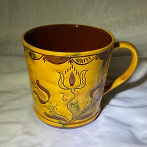 May include: A yellow ceramic mug with a brown rim and handle. The mug is decorated with a floral design in brown and green.