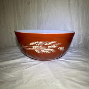 Puede incluir: Un cuenco mezclador vintage con un diseño de espigas de trigo. El cuenco es de color marrón rojizo cálido en el exterior y blanco en el interior. Las espigas de trigo son de color crema. Este cuenco es probablemente de mediados del siglo XX.
