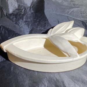 Puede incluir: Un macetero de cerámica blanco con forma de barco y un diseño floral. El macetero tiene un acabado liso y brillante y es perfecto para exhibir plantas o flores pequeñas.