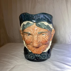 May include: A ceramic mug in the shape of an elderly person's face. The mug has a blue hat, a white collar, and a dark blue bow tie. The face is detailed with wrinkles and a smiling expression.