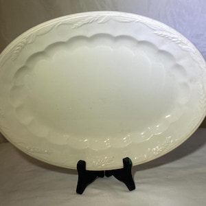 Puede incluir: Una fuente ovalada de color crema con un borde festoneado y detalles en relieve de trigo y flores. La fuente está sostenida por un soporte negro. Una pieza decorativa para servir alimentos.