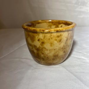 Puede incluir: Un pequeño cuenco de cerámica redondo con un patrón moteado y jaspeado en tonos beige y marrón. El borde es de un marrón más oscuro. El cuenco tiene un acabado liso y brillante y un diseño sencillo y clásico.