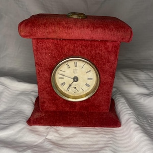 May include: A vintage red velvet clock. The clock face has Roman numerals and a second hand. The clock is rectangular with a small base and a decorative top. The clock face is round and set in a gold-coloured frame. The clock is likely from the early 20th century.