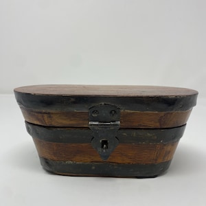 May include: A small, oval-shaped wooden box with a dark brown finish. The box is banded with black metal and features a metal clasp closure. The lid is also made of wood and has a dark brown finish. The box appears to be vintage or antique.