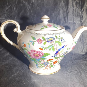 May include: A white teapot with a gold trim and floral design. The teapot features a bird and flowers painted on it.