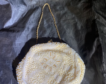 Puede incluir: Un bolso de noche blanco de cuentas con una correa de cadena dorada. El bolso es redondo y tiene un diseño floral.