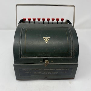 May include: Vintage dark green Todd Visible Adding Machine with red keys and a silver handle. The machine has gold accents and the text "Todd Visible Adding Machine" is visible on the front.