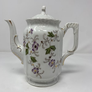 May include: A white porcelain teapot featuring a delicate floral pattern with purple flowers, green leaves, and gold-painted accents on the spout, handle, and lid. The teapot has a scalloped rim, a curved handle, and a matching removable lid.