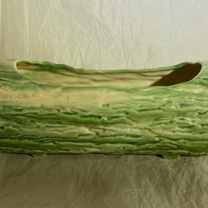 Puede incluir: Un macetero de cerámica verde con forma de tronco de árbol y textura de corteza rugosa. El macetero tiene una abertura rectangular para plantar.