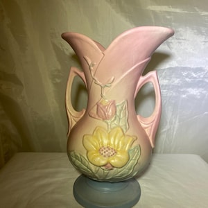 May include: A pink ceramic vase with a yellow flower and green leaves. The vase has a light blue base and handles. The flower is in the center of the vase. The vase is a decorative item.