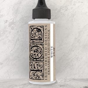 May include: A clear glass bottle with a black cap, containing white liquid chalk paint. The bottle is labeled "Iron Orchid Designs Erasable Liquid Chalk White 2 fl oz".