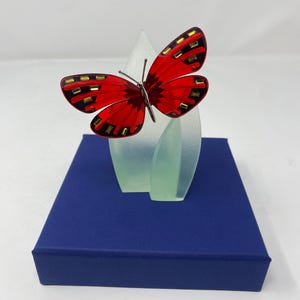 May include: A red butterfly figurine with black and gold accents perched on a translucent frosted green leaf-shaped base. The entire piece is mounted on a solid dark blue square pedestal, creating a contrast between the vibrant red wings and base.